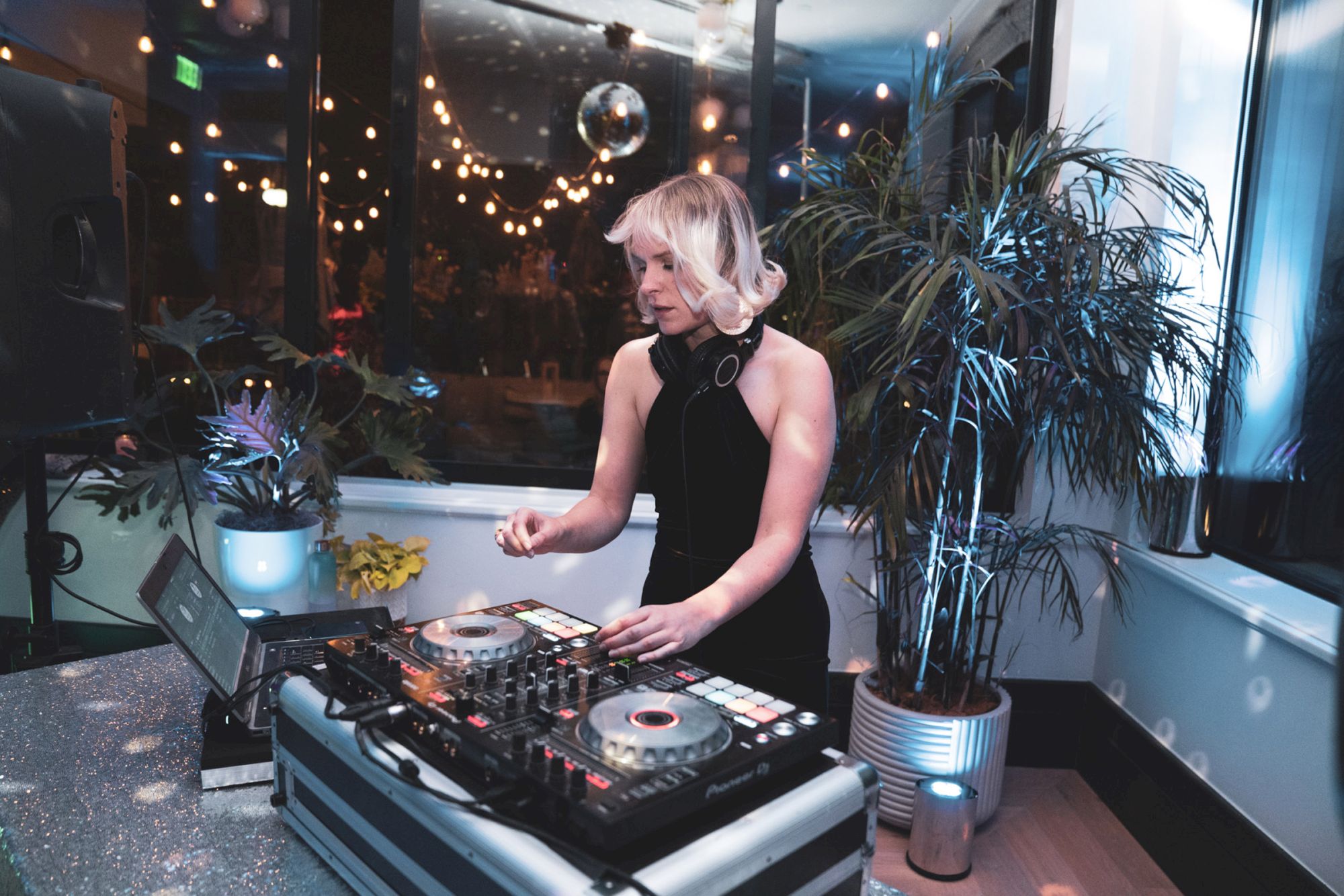 A DJ stands at a mixing console in a dimly lit room with plants and hanging lights, creating a lively atmosphere.