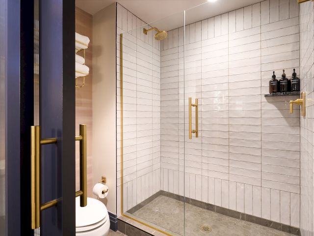 A modern bathroom with a glass shower enclosure, gold fixtures, white tiles, towels, and toiletries on a shelf is shown.