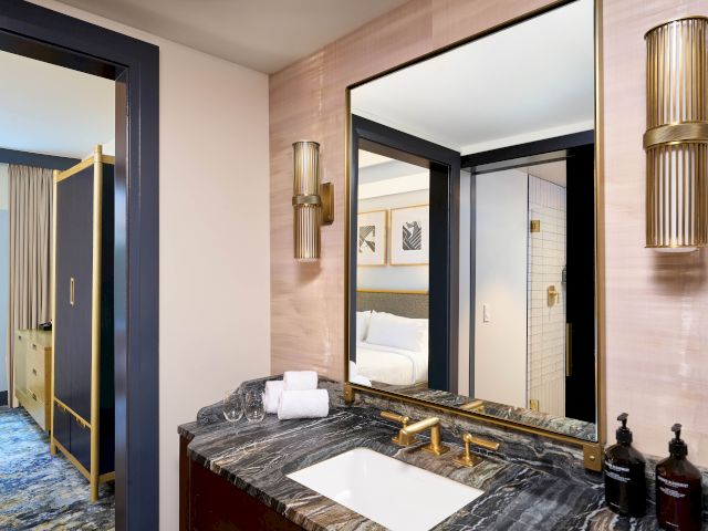 A modern bathroom with a marble sink, mirror, wall lights, and toiletries; a glimpse of a bedroom is visible through an open door.