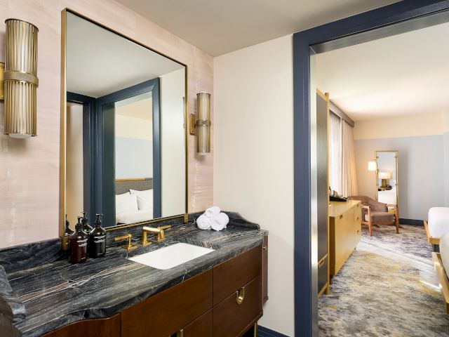 The image shows a modern bathroom with a marble-topped vanity, large mirror, and light fixtures, adjacent to a bedroom with a bed.