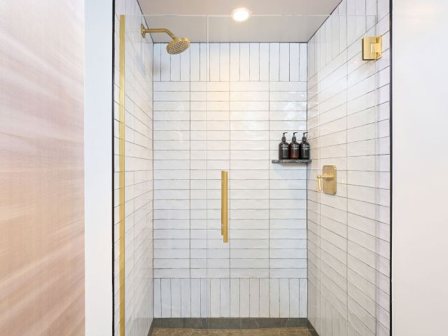 The image shows a modern shower with white subway tiles, a gold showerhead, and a small shelf holding bottles.