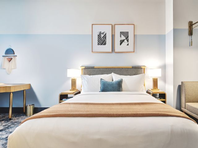 The image shows a hotel room with a bed, desk, and seating. It's decorated in neutral tones with wall art and stylish lighting fixtures.