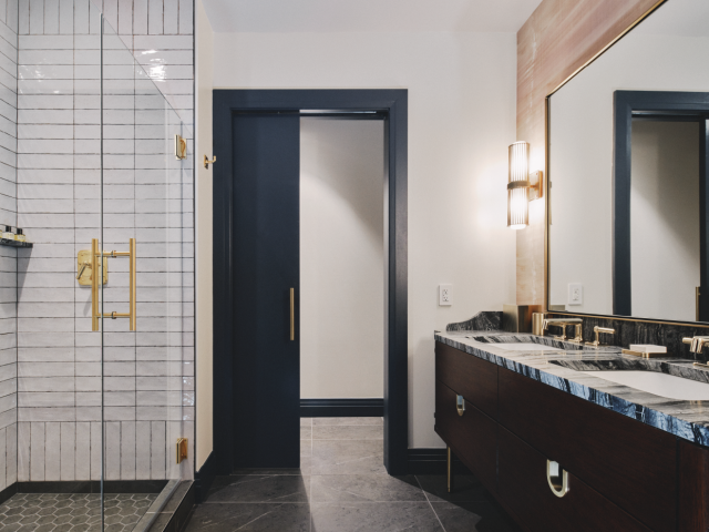 This image shows a modern bathroom with a glass shower, dark vanity, double sinks, and a large mirror under warm lighting fixtures.