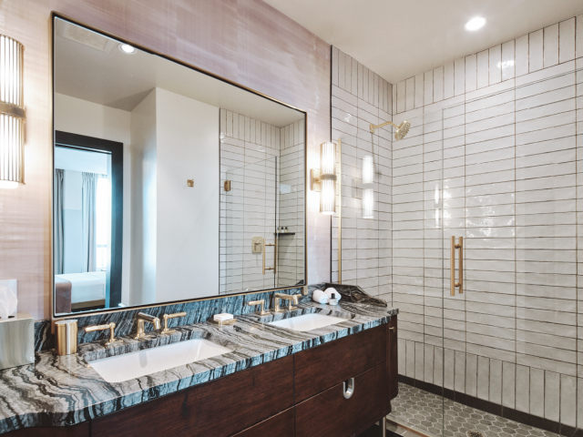 A modern bathroom features a large mirror, double sink with marble countertop, wall sconces, and a spacious shower area with a glass door.