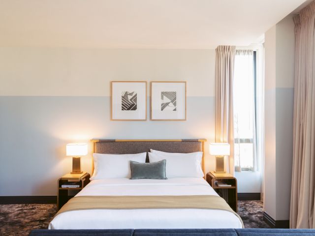 A neatly arranged hotel room with a double bed, two framed artworks above, and lamps on side tables next to a large window.