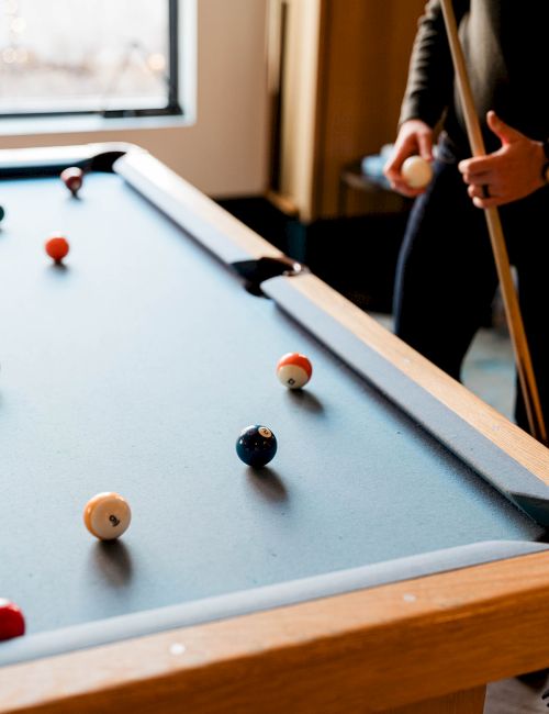 A person taps a pool cue on a blue-felt billiards table, with several balls scattered as they aim at a shot by the corner.