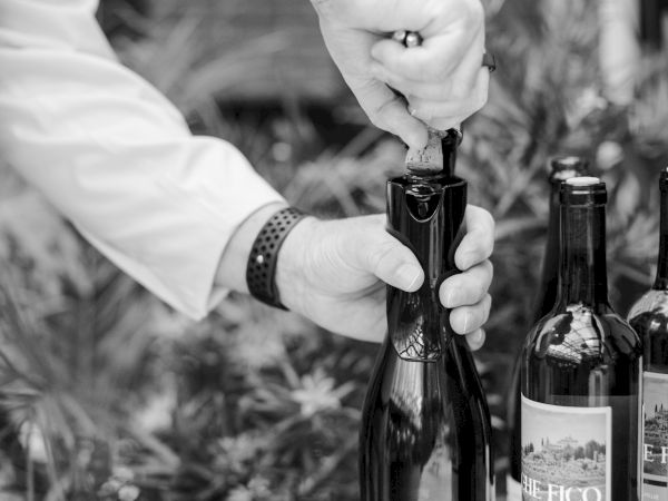 A person uncaps a wine bottle on a table outdoors, with two other bottles nearby, ready for pouring.