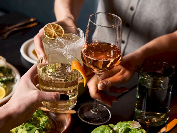 Friends clink glasses over a spread of salads and tapas, sharing drinks and laughter as a casual rooftop or restaurant meal vibe.