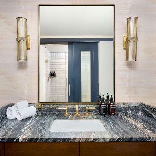 A bathroom vanity with a marble countertop, sink, and gold fixtures; mirror above, lights on sides, toiletries, and towels included.