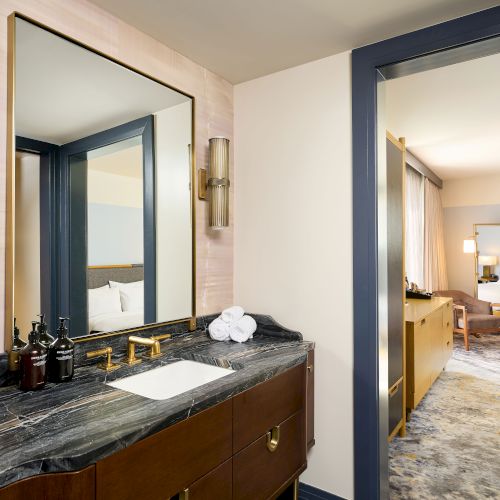 The image shows a modern hotel room with a bathroom vanity, mirror, and two beds visible through an open doorway.