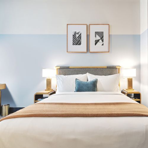 A stylish hotel room with a bed, side tables, and artwork on the wall, featuring modern lighting and a small sitting area with a desk.