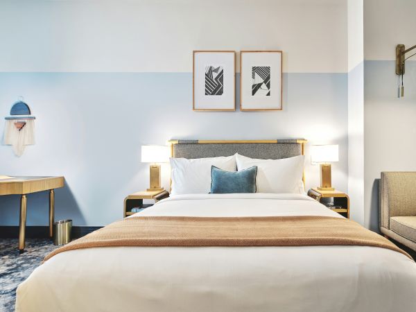 A modern hotel room with a queen bed, blue-gray color scheme, two nightstands, desk, chair, wall art, and a small seating area.