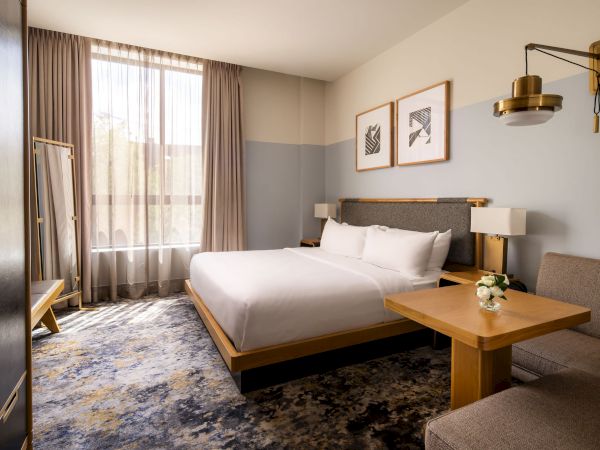 A modern hotel room with a queen bed, gray-blue accents, a wooden desk and bench, wall art, a large window with curtains, and a patterned rug.
