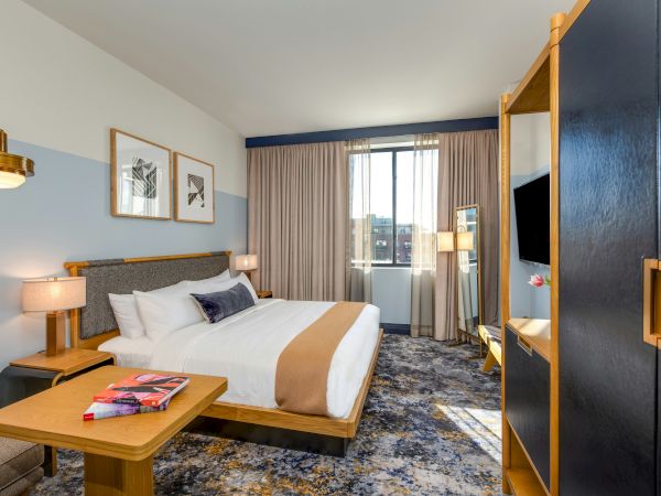 A modern hotel room with a wooden bed, white and beige bedding, two bedside lamps, a coffee table with magazines, a blue wardrobe, and a large window with beige curtains.