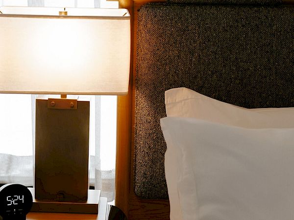 A neatly made hotel bed beside a nightstand with a lamp, a black hat, and a white-tiled window in the background.