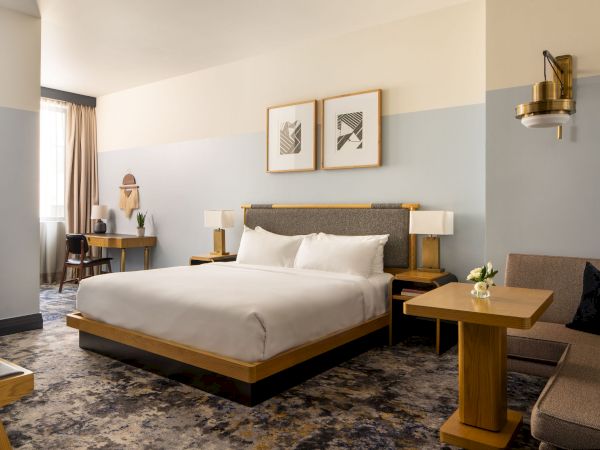 A modern hotel room with a king bed, soft blue wall accents, two nightstands, a desk, and a sofa; framed art above the bed and reading lamps.