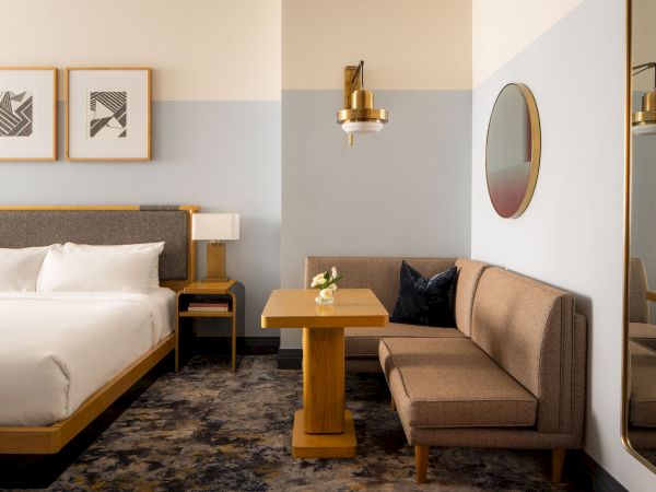 A modern hotel room with a king bed, a small seating nook, a wooden table, framed art, round mirror, and warm pendant lights, tidy and inviting.