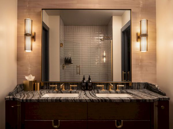 A luxurious powder room with a double sink, marble countertop, wood vanity, large framed mirror, and modern sconces on either side, reflecting a tiled shower behind.