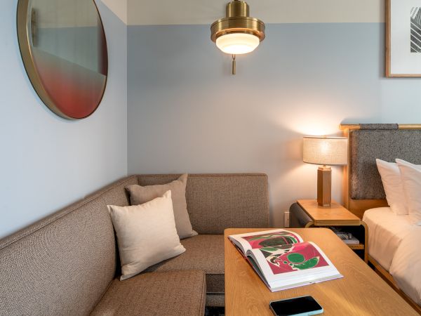 A cozy hotel room with a beige sofa, wooden coffee table, bed, lamp, artwork, and a round mirror&mdash;perfect for a relaxing stay.