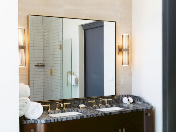 A modern bathroom vanity with a double sink, marble countertop, large mirror, wall sconces, towel rolls, and a glass shower in the background, chic and bright.