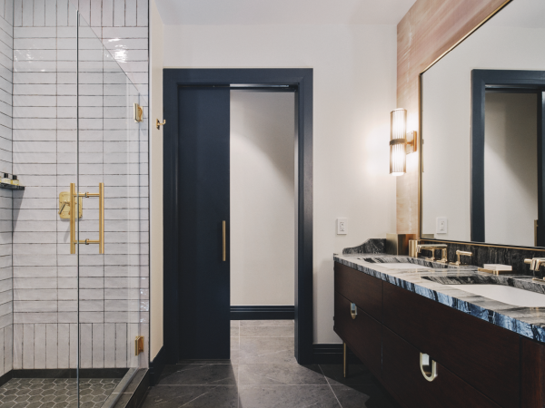 A modern bathroom with a glass shower, double sink vanity, and dark accents, featuring gold fixtures and a large mirror on the wall.
