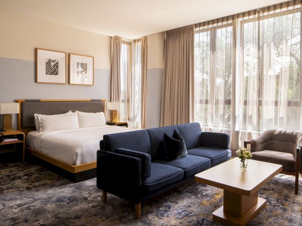 A modern hotel suite with a bed, two nightstands, a navy sofa, a coffee table, an armchair, and large windows with sheer curtains, greeting natural light.