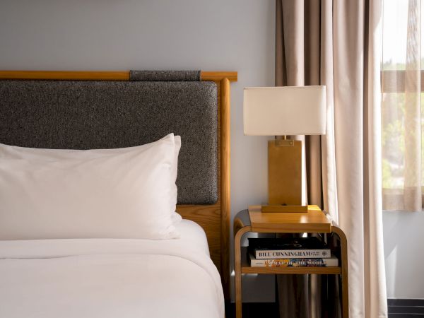 A neatly made hotel bed with white linens and a padded headboard beside a bedside table holding a lamp and magazines, near a window with curtains.