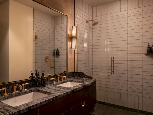 A luxurious bathroom with a double sink, dark marble countertop, gold fixtures, a large mirror, and a glass-enclosed shower with white subway tiles.