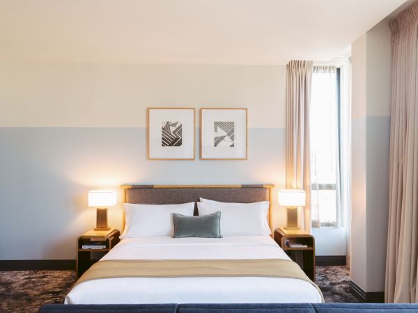 The image shows a neatly arranged hotel room with a bed, two side tables with lamps, artwork above the bed, and a window with curtains.