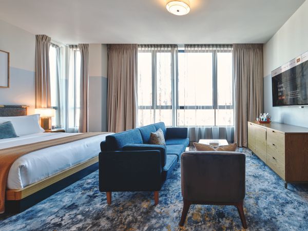 A spacious modern hotel room with a king bed, blue accents, seating area, and a flat-screen TV, plus large windows and soft lighting.