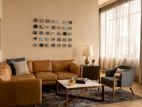 A cozy living room with a tan leather sofa, a matching armchair, a wooden coffee table, a striped rug, and wall art made of small square panels, near a large window.