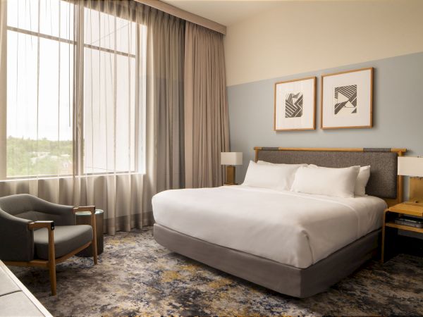 A modern hotel room with a large bed, gray headboard, two bedside tables, a chair by the window, and framed wall art above the bed.