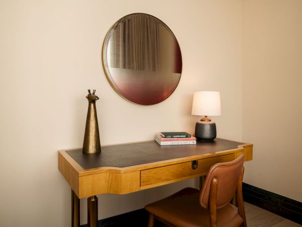 A wooden desk with a black top, a round wall mirror, a small table lamp, a few books, and a brown chair in a minimalist room.