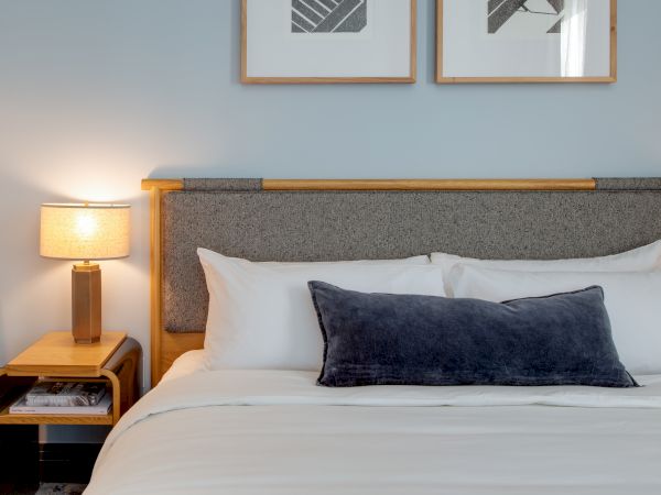 A neatly made hotel bed with white sheets, a dark throw pillow, bedside lamp, and framed abstract art above the headboard.