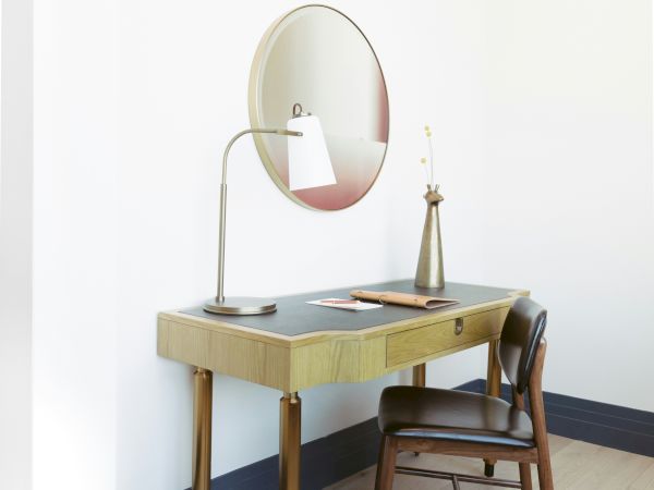 A minimalist wood vanity with a round mirror, a desk lamp, and a wooden chair sits against a pale wall in a bright, airy room.