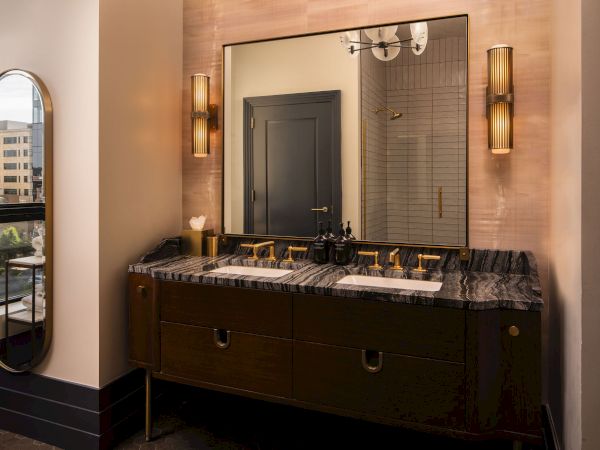 A stylish bathroom with a double sink vanity, granite countertop, large mirror, wall sconces, and a slim arched mirror reflecting a city view, ending with a period.