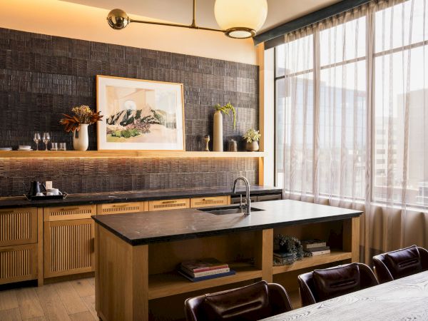 A modern kitchen with a central island, dark tiled backsplash, wooden cabinets, and large windows letting in natural light, plus bar stools along a counter.