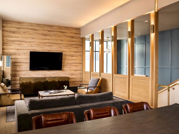 A modern, cozy living room with a wall-mounted TV, wood-paneled feature wall, gray sofas, and warm lighting; glass partitions and a high-end dining area nearby.