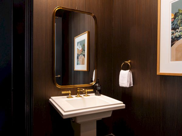 A small bathroom with a pedestal sink, brass faucet, wall-mounted towel ring, dark wood paneling, a rectangular mirror, and framed art on the wall.