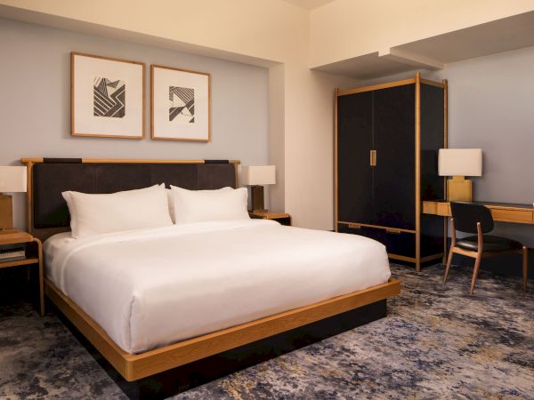 A modern hotel room with a large bed, two nightstands, framed art above the headboard, a wardrobe, desk with chair, and a softly lit ambience.