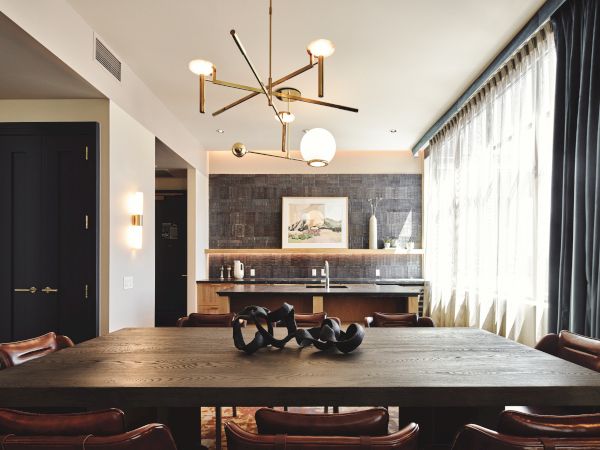 A modern dining room with a large wooden table, stylish chandelier, dark chalkboard wall, bar area, and floor-to-ceiling windows with flowing curtains.