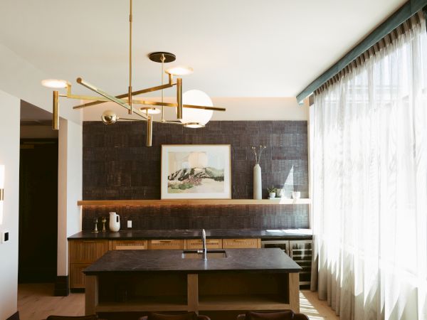 A modern kitchen with dark cabinets, a central island, pendant lights, wall art, and large window curtains allowing natural light in.