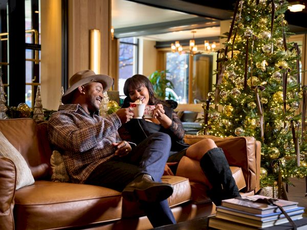 A couple relax on a couch at home, sipping hot drinks and enjoying a Christmas tree lit with golden lights and ornaments.