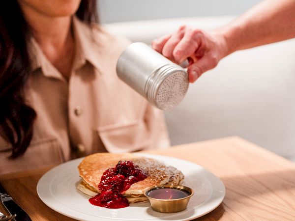 A person smiles as a server sprinkles powdered spice over a plated pancake with berry compote and sauce on a table.