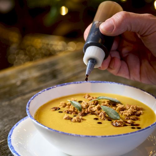 A hand drizzle-decorates a creamy orange soup with a squeeze bottle, sprinkled with nuts and mint in a blue-rimmed bowl, cozy lighting.