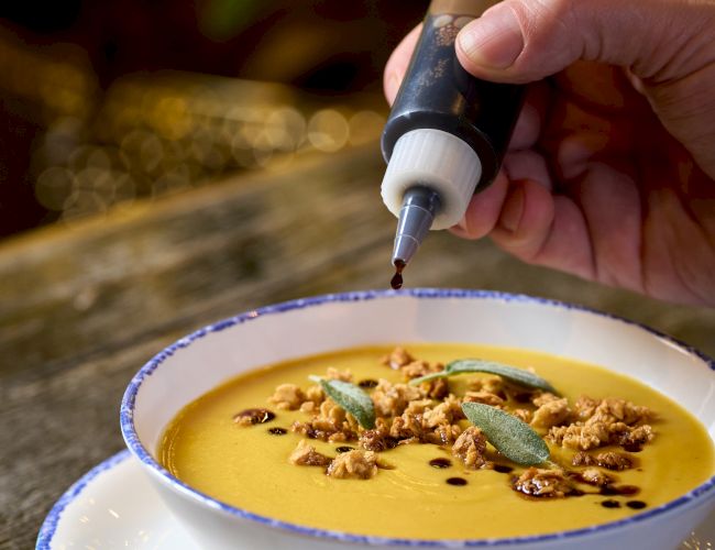 A hand drizzle-decorates a creamy orange soup with a squeeze bottle, sprinkled with nuts and mint in a blue-rimmed bowl, cozy lighting.