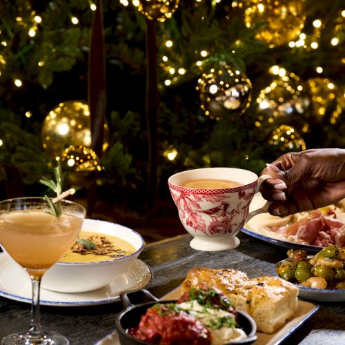 A festive table with cocktails, tea, and assorted tapas in front of glowing string lights, creating a warm, elegant mood.