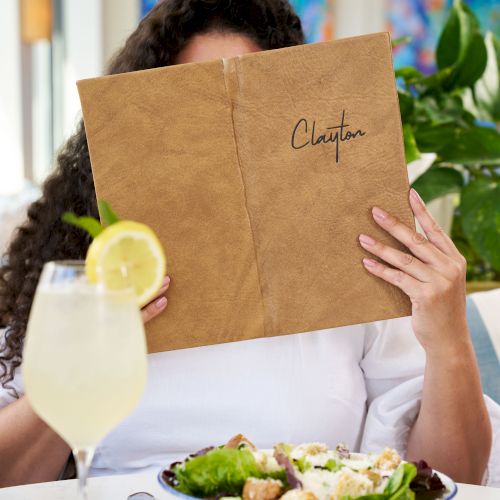 A person at a table reading a brown notebook titled &ldquo;Chapter,&rdquo; with a salad, a lemon drink, and a blurred colorful background.