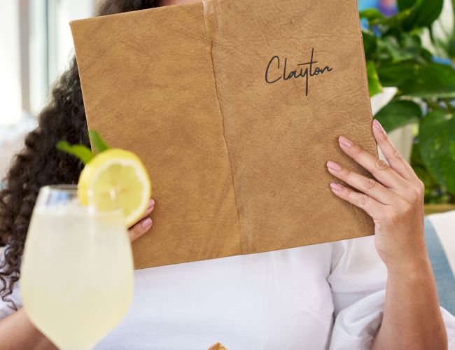 A person at a table reading a brown notebook titled &ldquo;Chapter,&rdquo; with a salad, a lemon drink, and a blurred colorful background.