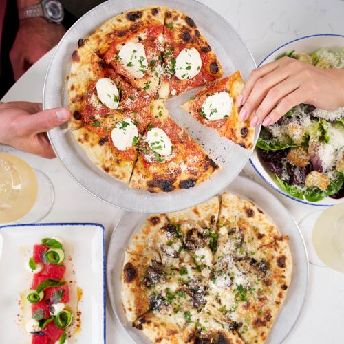 A table of pizzas and salads with lemon drinks; hands share a slice while toppings include mozzarella and herbs, delicious vibes.